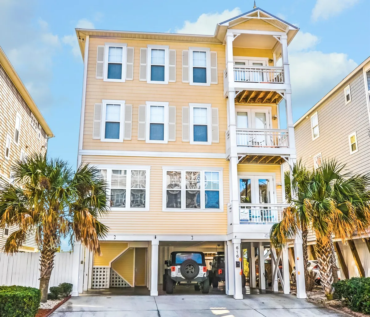 The exterior of a three-story beachfront building with multiple balconies, surrounded by palm trees and a driveway for easy arrivals. The image conveys a welcoming, family-friendly atmosphere and highlights the building's proximity to the beach.