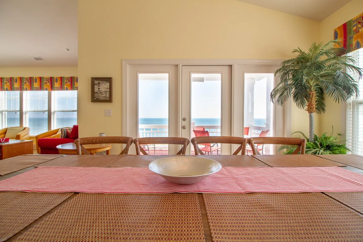A bright dining area with a large table set near expansive windows that reveal a stunning ocean view. The space is decorated with a palm plant and colorful curtains, giving it a welcoming, coastal feel. Natural sunlight pours in, illuminating the area where families can gather for meals and enjoy the scenery just beyond the glass.