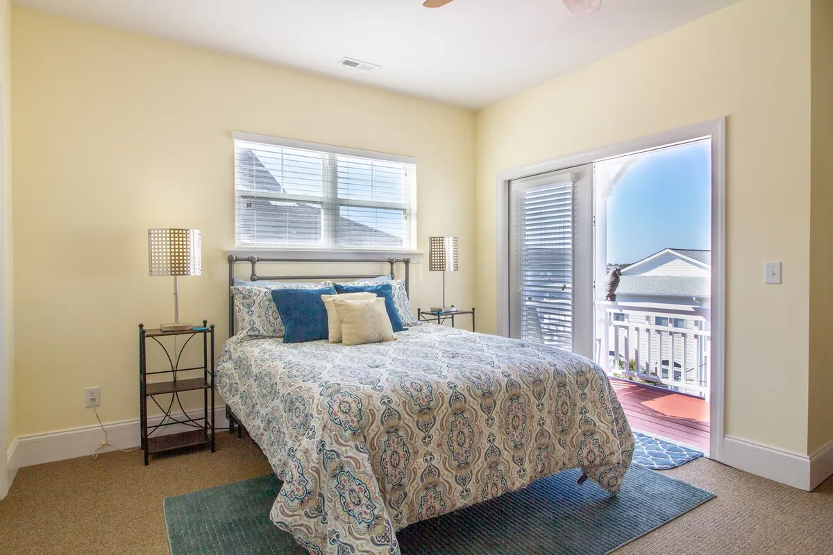 A spacious bedroom featuring a bed with patterned bedding, decorative pillows, nightstands with lamps, and an open door leading out to a balcony. Sunlight streams into the room, and the balcony offers a glimpse of the outdoors, suggesting access to fresh air and ocean views.
