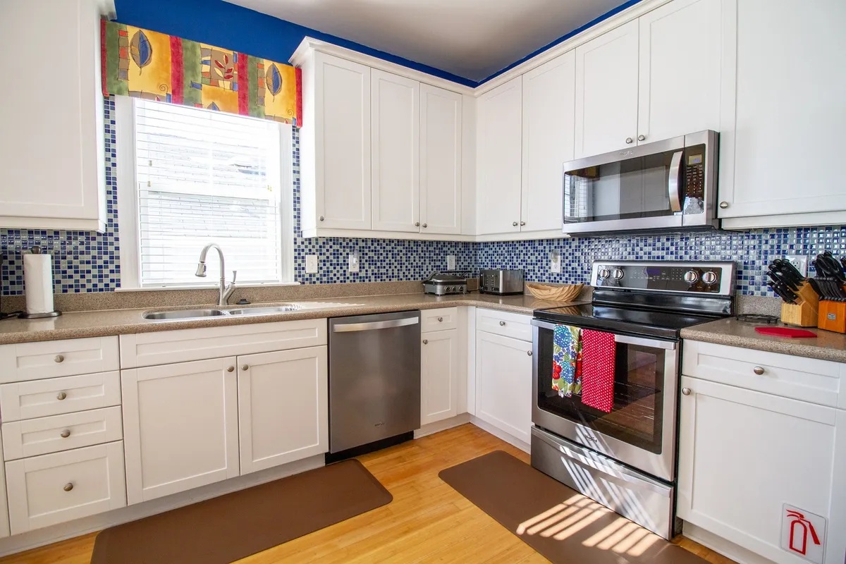 A welcoming view of the kitchen space featuring a colorful window treatment, mosaic backsplash, stainless steel appliances, and warm wooden flooring. The layout encourages both meal prep and group conversation.