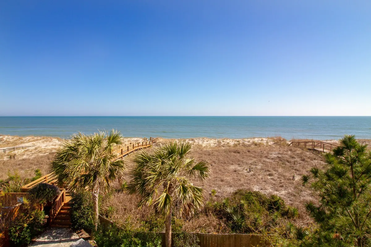 A sweeping view of the sandy beach and Atlantic Ocean, framed by palm trees and a private boardwalk leading directly from the property to the shore. The scene captures clear skies, inviting sand, and the gentle waves, giving a sense of immediate and effortless beach access.
