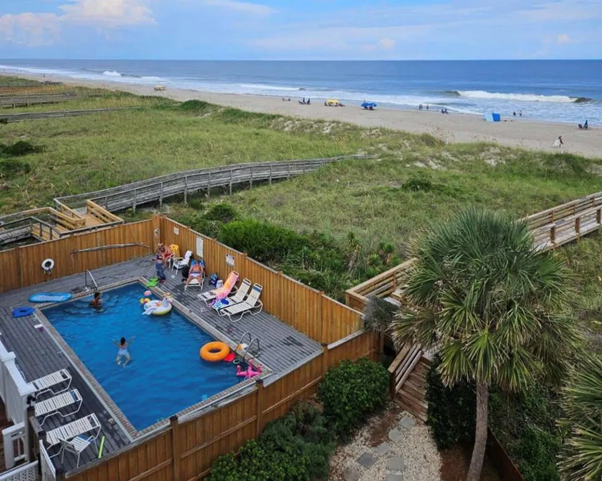 This vibrant exterior image captures the shared oceanfront swimming pool, with guests lounging on chairs, people swimming, and the beach visible just beyond. Lush palm trees and poolside decorations contribute to the atmosphere of leisure and fun, emphasizing the direct connection between poolside enjoyment and immediate beach access.