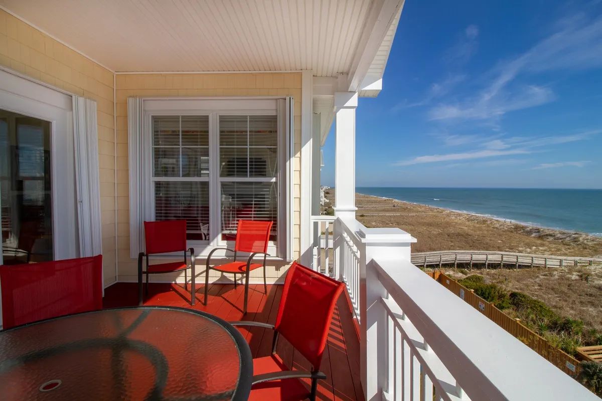 An exterior shot of a covered oceanfront deck furnished with red patio chairs and a glass table. The view captures the rolling waves and sunrise or sunset over the Atlantic, inviting guests to relax, sip coffee, or dine al fresco while basking in the seaside ambiance.