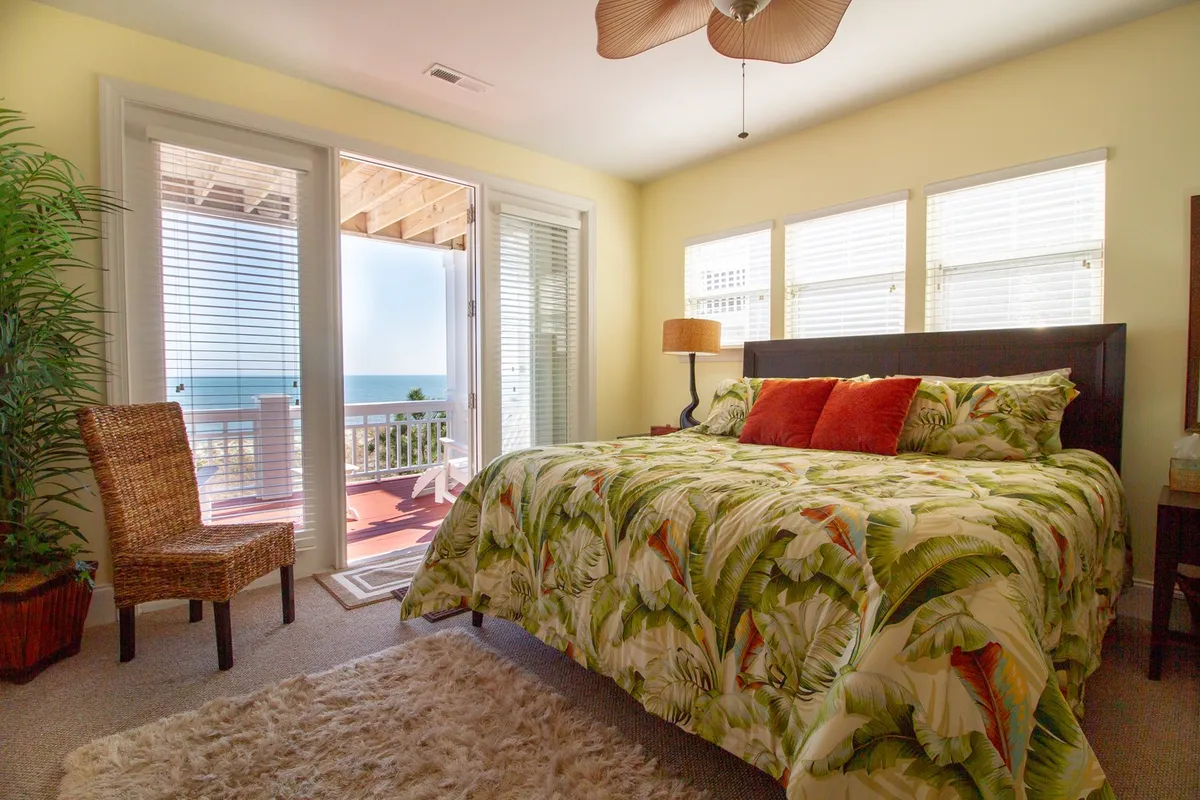 A tranquil bedroom with a large bed adorned with tropical-themed bedding, comfortable seating, decorative lamp, and open doors revealing a stunning sea view. The room captures a relaxing, retreat-like ambiance.