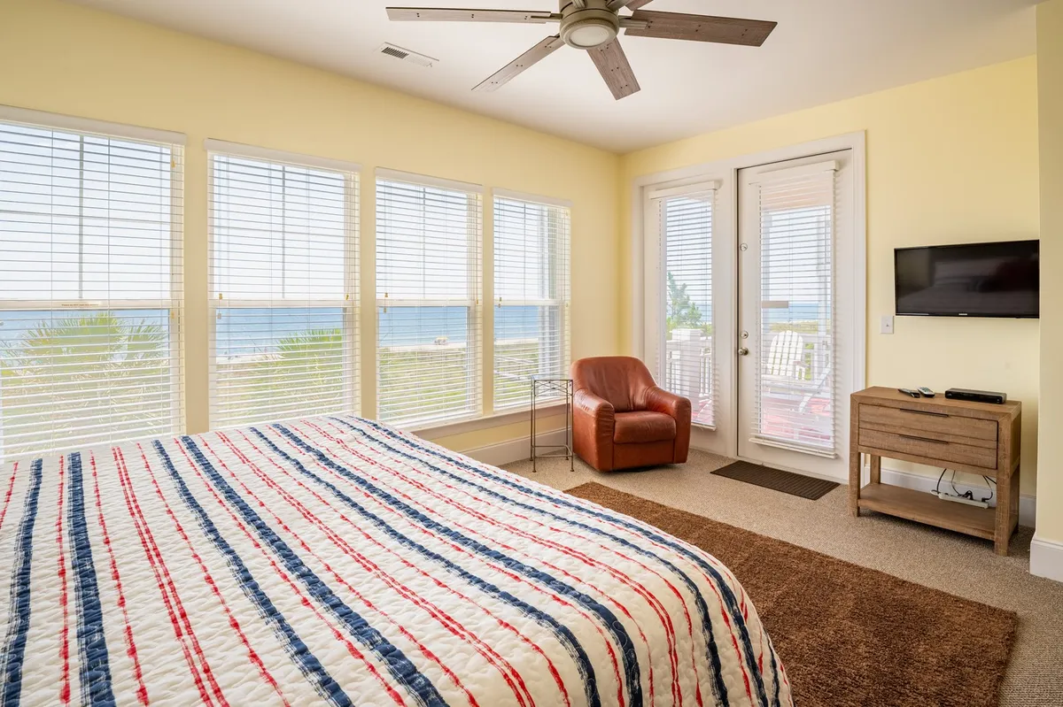 A serene, sun-filled bedroom featuring a bed with a striped quilt, large windows framing panoramic ocean views, a red armchair for reading, and a wooden dresser. The space exudes calm with its soft natural light and inviting textures.