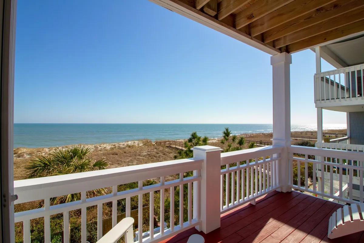 An elevated oceanfront balcony view with a wooden railing overlooking the beach and lush natural vegetation. The wide-open perspective showcases the blue sky, rolling waves, and tranquil setting—ideal for watching family play or enjoying sunsets together.