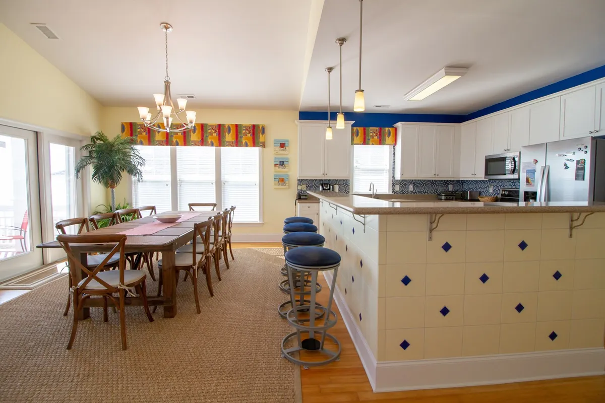 A spacious, open dining area next to a modern kitchen island, with a welcoming chandelier overhead and window views that suggest a bright, coastal setting. The space is set up for meals, cooking, and lively conversation.