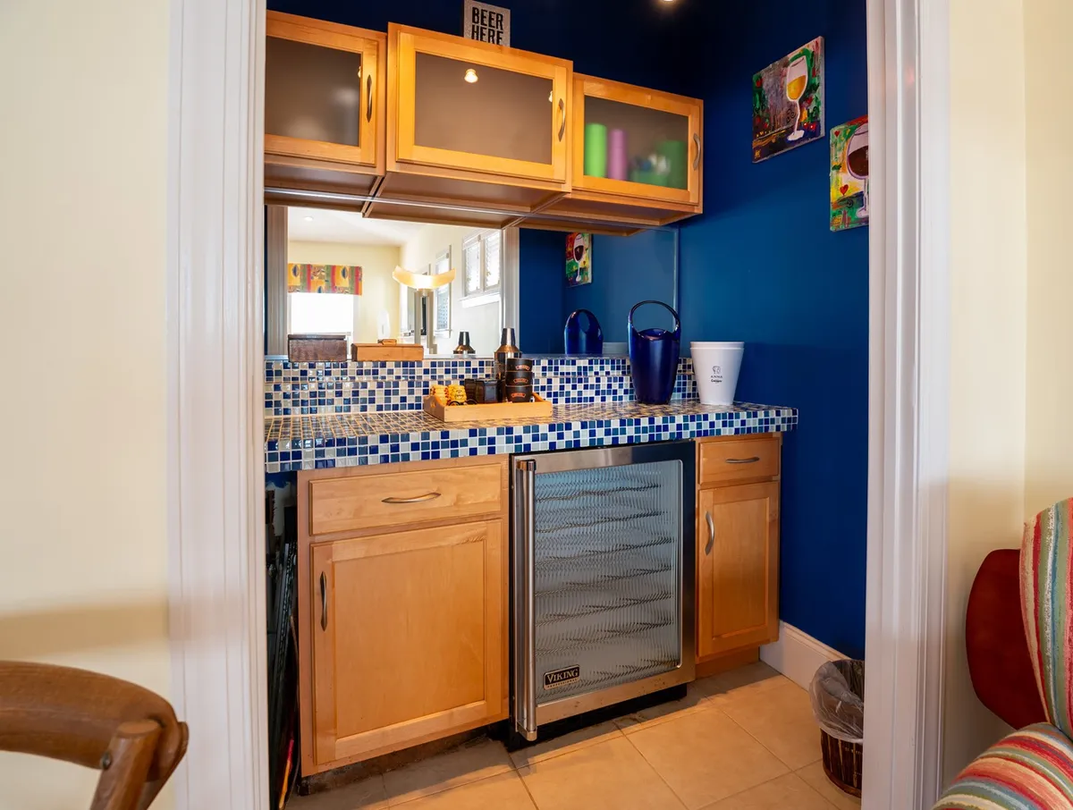 A lively wet bar area with a colorful mosaic counter, blue accent wall with artwork, and an under-counter refrigerator (wine fridge). The space feels energetic and social—designed for preparing drinks and hosting festive gatherings.