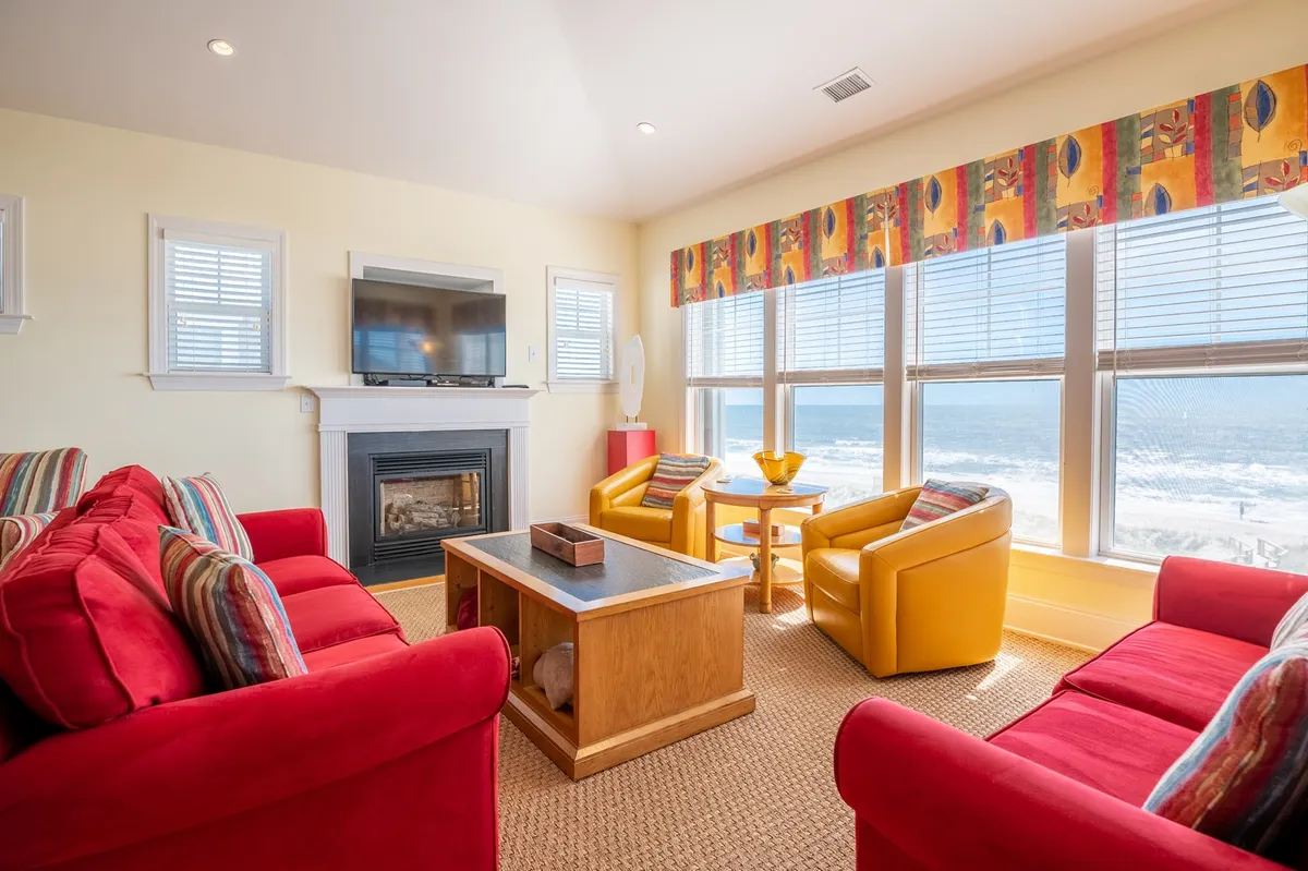 A cheerful, inviting living room featuring red sofas, yellow chairs, a coffee table, and a fireplace. Large windows overlook the ocean, filling the space with natural light and offering a stunning water view. The layout is open and perfect for gatherings and relaxation with family or friends.