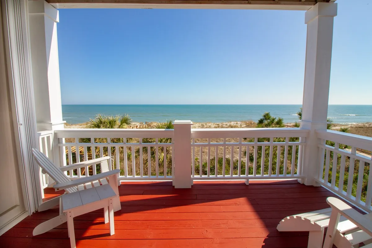 A private red wooden deck with white railings, furnished with classic Adirondack chairs, overlooks a stunning ocean vista. The scene blends indoor-outdoor living and is perfect for quiet reflection, reading, or stargazing.