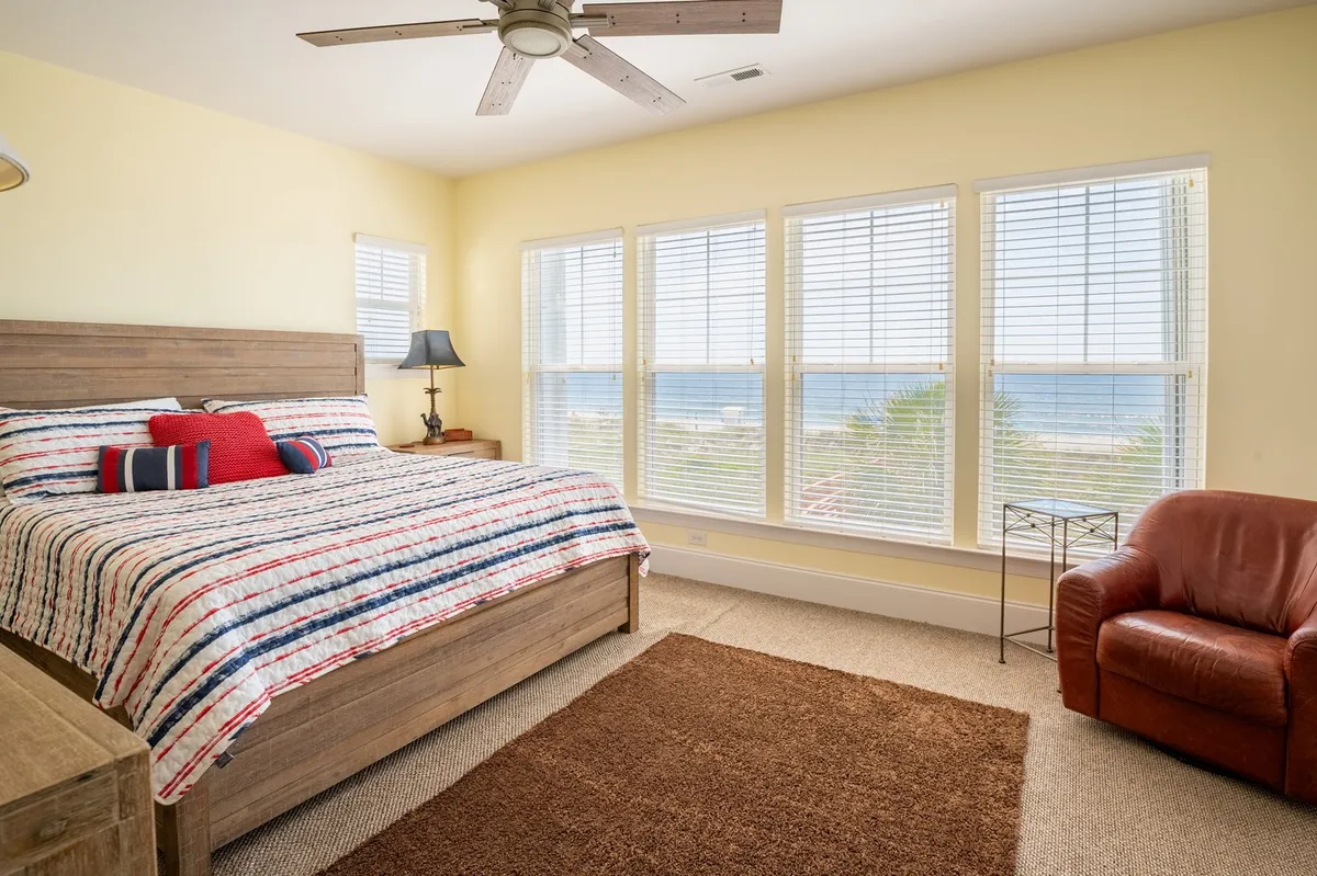 A serene bedroom with colorful bedding, a leather armchair, and large windows that showcase panoramic ocean views. The space is inviting and tranquil, ideal for adults or grandparents to relax, unwind, and enjoy the sights and sounds of the coast.