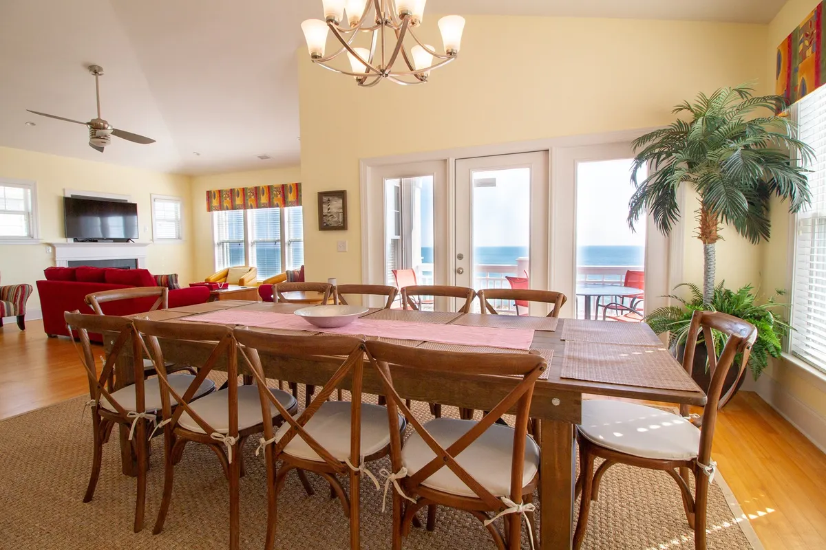 A spacious dining area with a large wooden table that comfortably seats ten beneath a glowing chandelier. Glass doors in the background open to a view of the ocean, and a tropical plant adds a fresh touch. The setting is ideal for celebratory dinners or casual brunches, with natural light enhancing the welcoming ambience.
