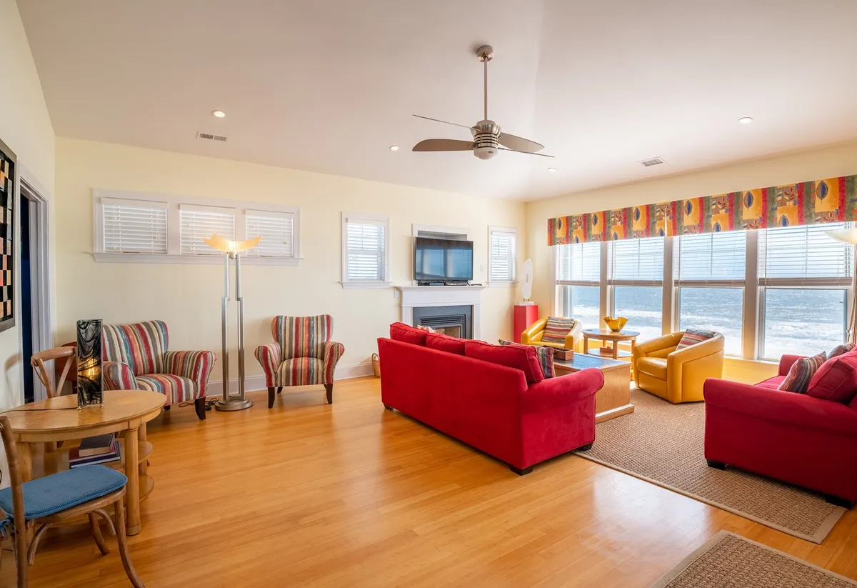 A bright, airy living space featuring vibrant red sofas, large windows with lots of natural light, colorful curtains, a wooden coffee table, and striped armchairs. The view through the windows hints at the proximity to the beach and outdoor activities.