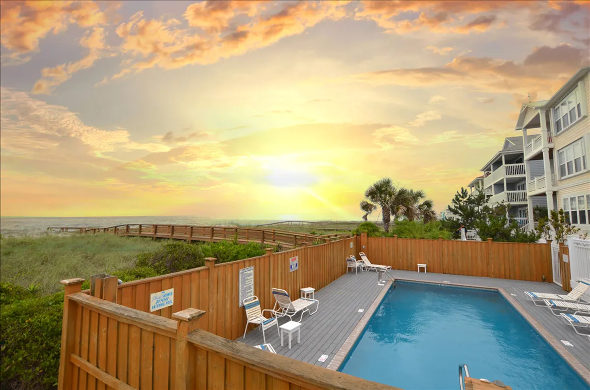 A serene view of the swimming pool area at sunset, framed by a wooden deck and swaying palm trees. The pool glistens in the evening light, inviting guests for a peaceful swim or relaxed gathering after a day at the beach.