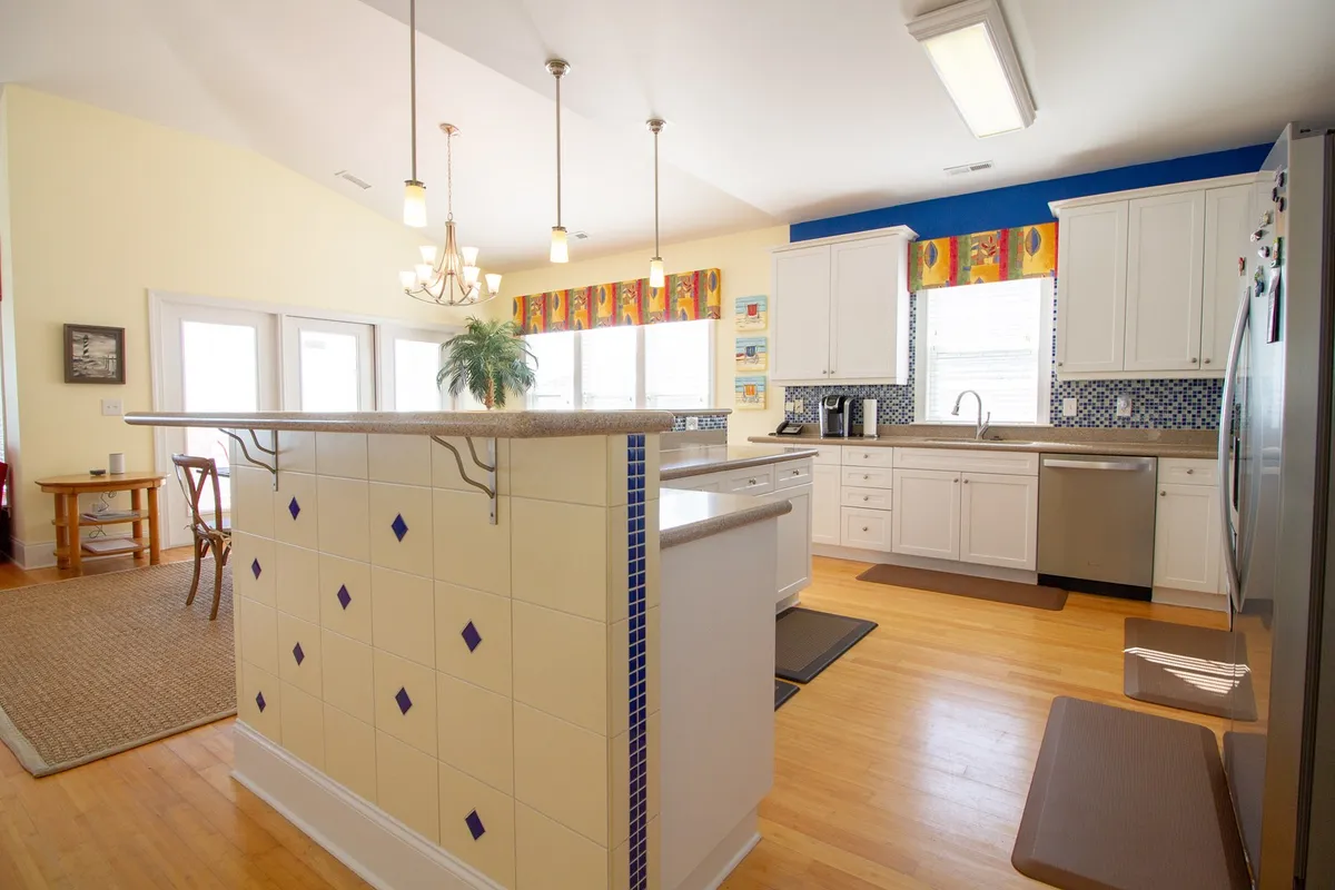 A spacious kitchen with a large island featuring bar seating for four. The area is filled with natural light from large windows adorned with colorful window treatments. Modern appliances complete the scene, inviting both cooking and group gatherings.