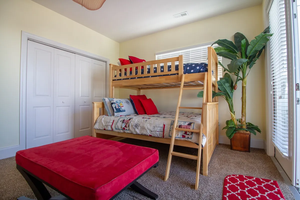 A cheerful bunk room featuring a sturdy bunk bed, a red ottoman, and a large potted plant. The room is designed to be playful and inviting, making it perfect for kids to sleep, read, and play. The bright window adds natural light and a sense of connection to the outdoors.