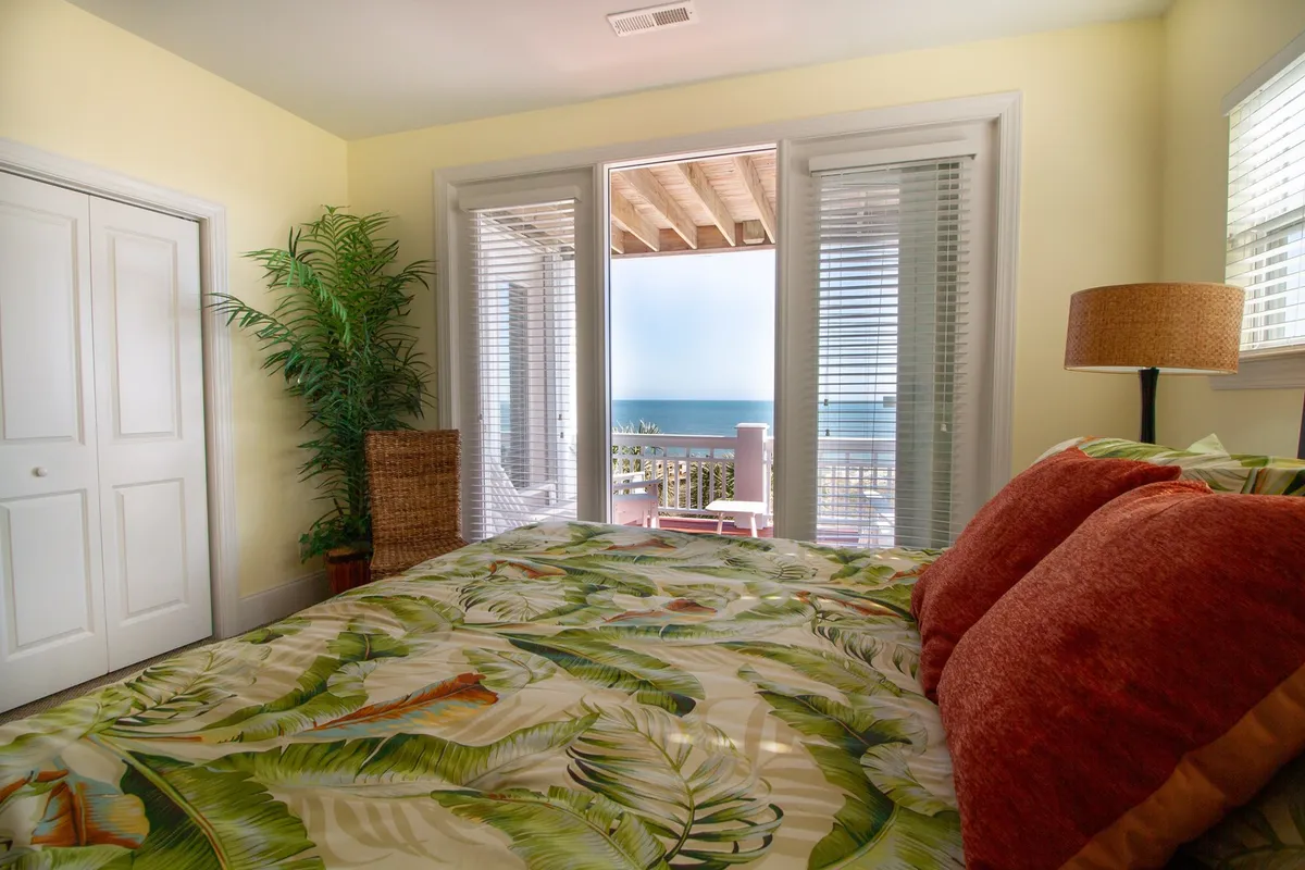 A serene bedroom with tropical-themed bedding, a decorative lamp, and large windows that offer sweeping ocean views. The peaceful atmosphere invites relaxation and indulgence in the coastal environment.